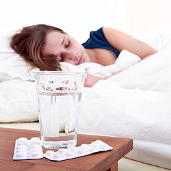 Glass of water and two strips of pills on a bedside table, with a sick woman sleeping in the background